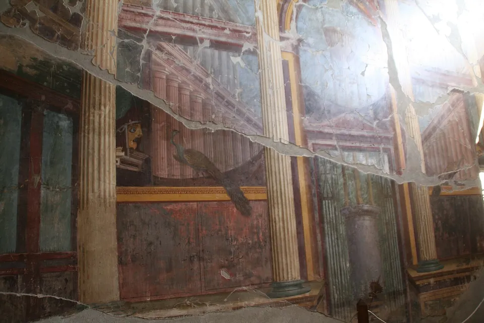 A cracked wall painting of fluted columns and receding architecture in red, ochre and blue-green, with a peacock standing on a ledge and a theatre mask to its left; light glares off the surface.