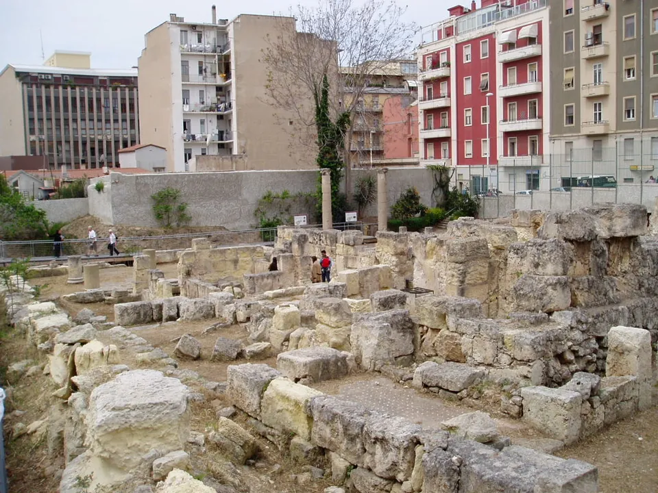 Low walls of squared limestone blocks marking out rooms on open ground, with a few standing columns and paths between them, a few visitors walking across the far side, and blocks of modern flats around the site.