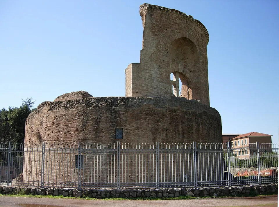 The lower drum of a round brick building, its top rough and broken, with a tall curved section of the upper storey standing above it, pierced by arched openings, behind an iron fence under a clear sky.