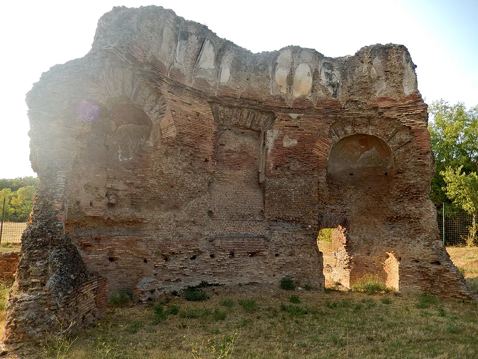A high, roofless brick wall with arched niches and a doorway opening through it, its top ragged, standing on grass in low evening sun.