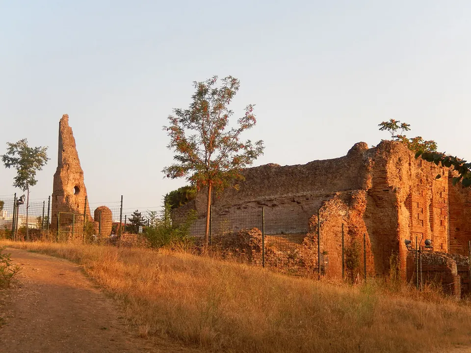 A long ruined brick building behind a wire fence beside a dirt path through dry grass, with a tall thin ruin rising in the distance on the left.