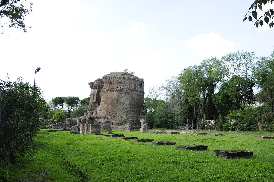 A broad mown lawn crossed by two rows of low, squared stone bases, leading towards a round brick ruin with a broken top standing among trees under a pale sky.