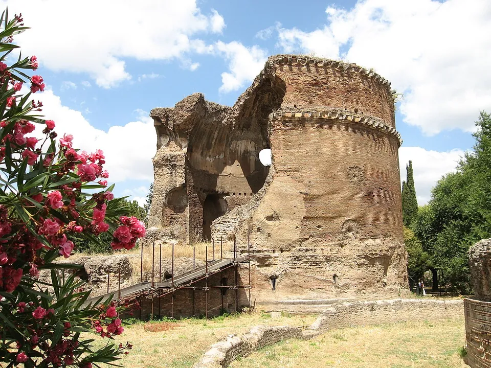 A tall cylindrical brick building with two projecting cornice bands, its upper part broken open on one side to show the curve of a dome and a round window, with a metal stair against its base and oleander flowers in the foreground.