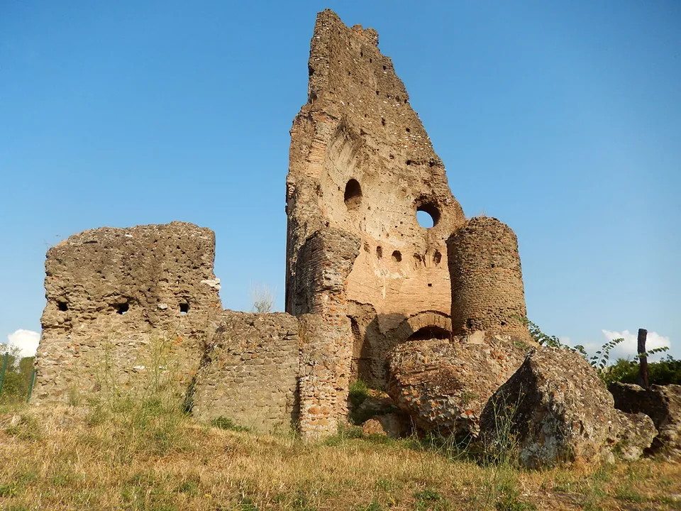 A tall, jagged fragment of brick and concrete wall pierced by round holes and a round window, with a smaller round turret and low broken walls at its foot, against a blue sky.