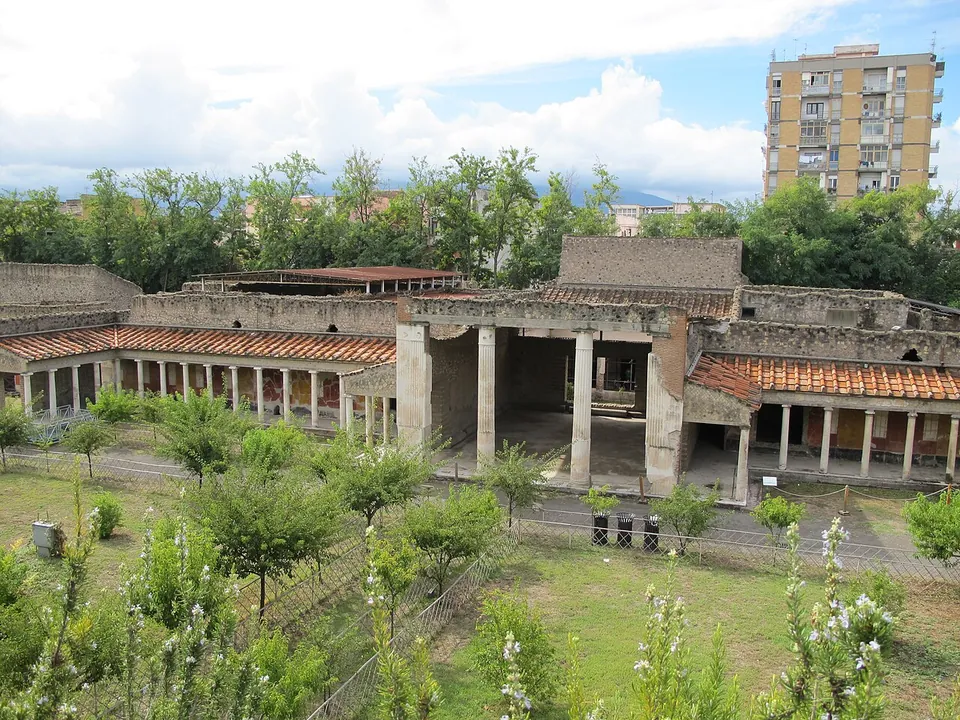 A long front of rough stone walls with porticoes of slender columns under red-tiled roofs and a tall square opening between heavy piers in the middle, seen across a lawn with young trees and flowering shrubs, with a modern apartment block rising behind.