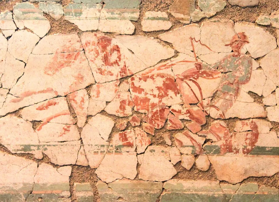 A wall painting pieced together from many cracked fragments, showing in red outline a charioteer in a helmet driving horses at speed, above a green band.