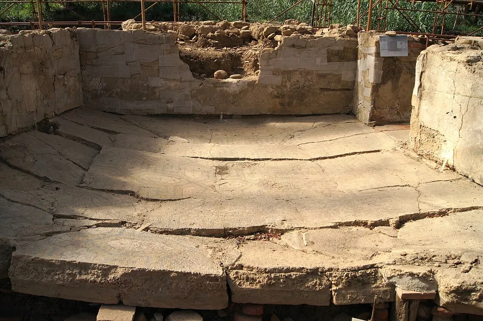 A sunken room floor of pale tesserae, its faint geometric pattern crossed by long cracks where the floor has sagged and split, enclosed on three sides by low walls of squared sandstone blocks with traces of plaster, under a frame of scaffolding poles.