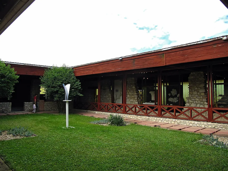Low museum buildings with dark red timber roofs and railings around a lawn, with rough stone walls visible behind their glazing and a slender white sculpture on the grass.