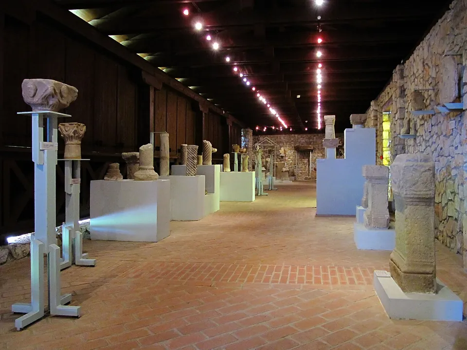 A long hall with a brick floor and stone walls, lined with carved stone blocks, capitals and altars on white plinths under spotlights.