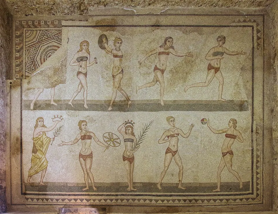 A floor mosaic in two rows of young women in two-piece athletic dress, one lifting small weights, others running, throwing a discus and passing a ball, with a robed figure in the lower row holding out a crown and a palm branch.