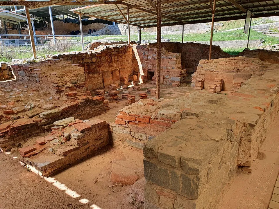 Low walls of squared grey stone and red brick divide a sandy floor into small rooms beneath a metal shelter held up on steel posts.