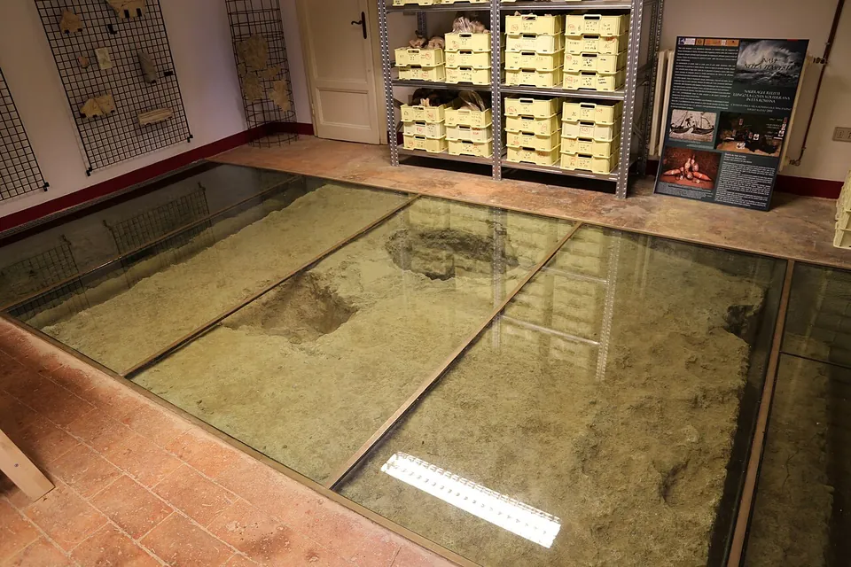 A room lined with shelves of stacked plastic crates, part of its tiled floor replaced by glass panels through which a curved surface of earth and masonry is visible below.