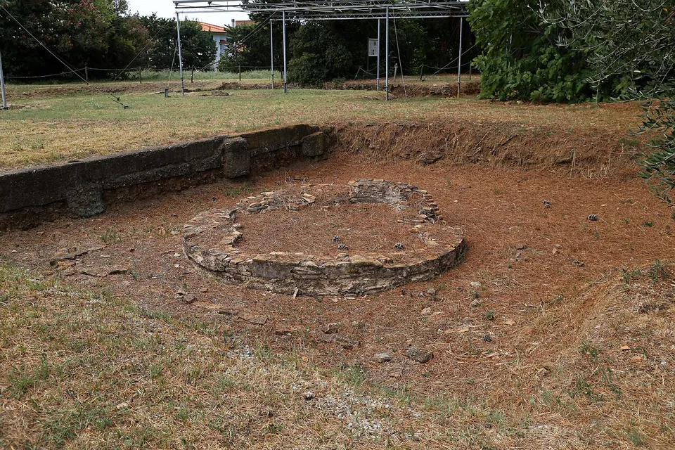A low ring of stone, the base of a round fountain, sits in a shallow sunken area of bare earth, with a low stone retaining wall behind it and, beyond, grass, trees and the metal frames of protective shelters.