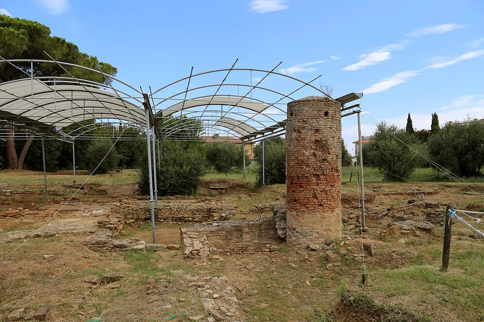 Low walls of rough stone spread across a dry grassy excavation under arched metal shelter roofs, with a round pier of red brick standing among them.