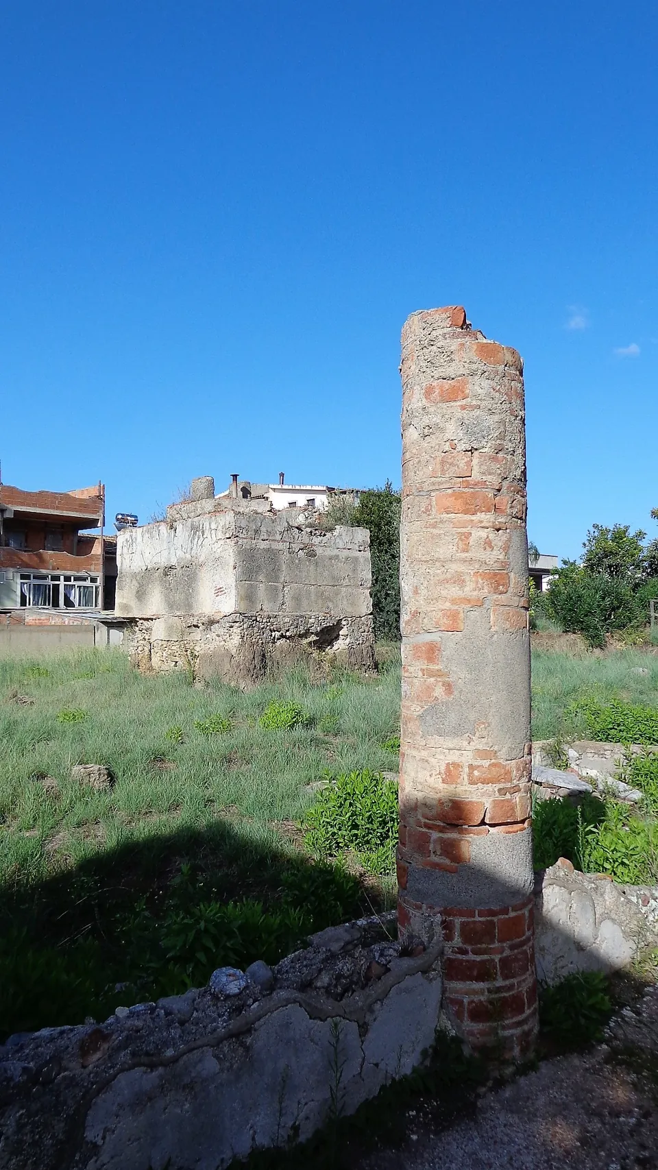 A single column built of curved bricks rising from low stone walls, with grass around it, a squared block of masonry behind and modern houses at the edge of the site under a blue sky.
