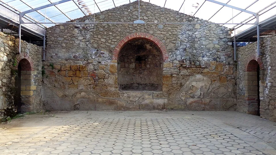 A large room under a modern glazed roof on a metal frame, its walls of rubble stone with a brick-arched niche in the end wall and an arched doorway on each side, the floor laid with hexagonal tiles.