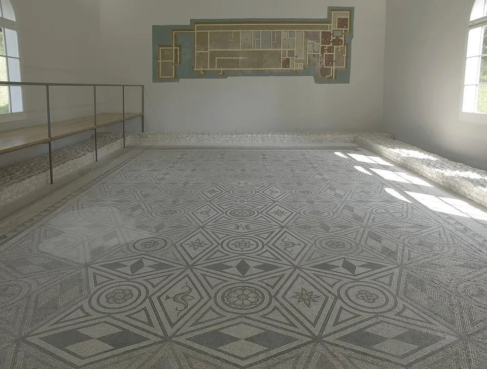 A large mosaic floor of black and white geometric patterns, stars and rosettes filling a bright room with arched windows, a railing along one side and a framed building plan on the wall.