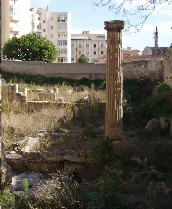 A single tall stone column standing among low ruined walls overgrown with dry grass and shrubs, with a high boundary wall and apartment buildings behind.