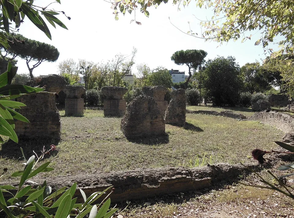Several rough stumps of rubble masonry standing in dry grass in a line, with a low wall in the foreground and pines and trees behind.