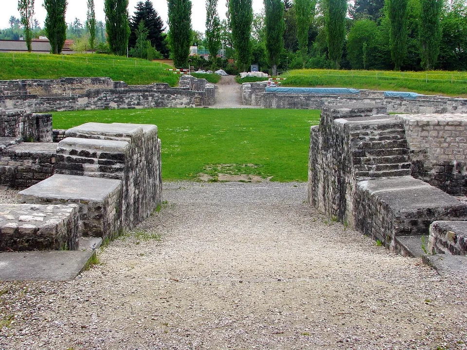 A gravel passage between massive walls of squared stone leading into a grassy oval arena, with more walls and a grass bank on the far side and a row of poplars behind.