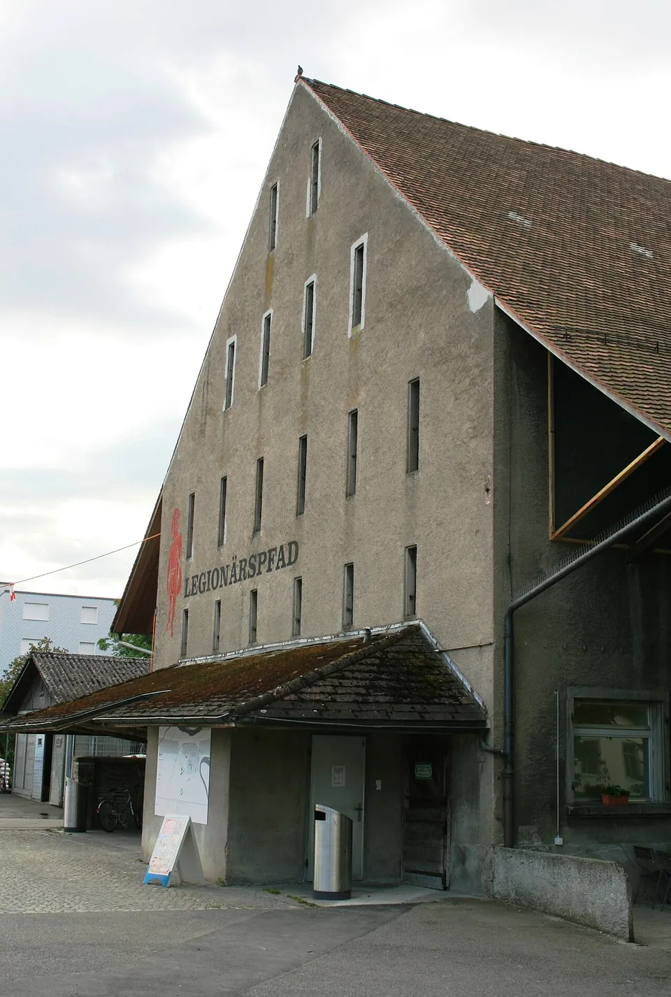 The tall gabled end of an old plastered barn with rows of small windows, the word LEGIONÄRSPFAD painted on it and a low entrance porch at its foot.