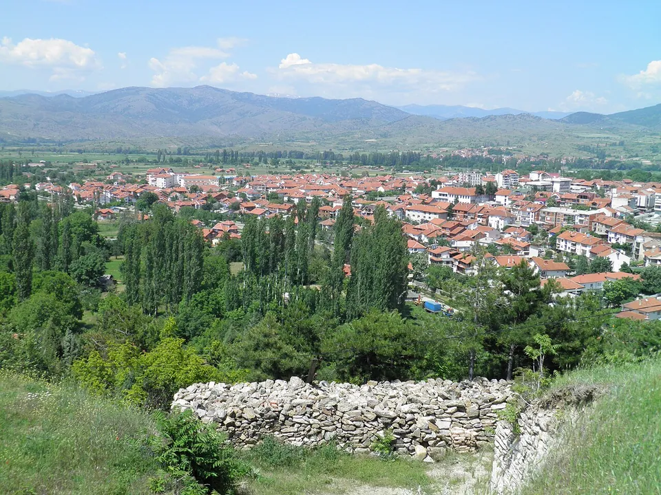 A stretch of dry-stone wall at the edge of a grassy slope, with poplars and the town of Vinica spread across the valley below and mountains on the horizon.