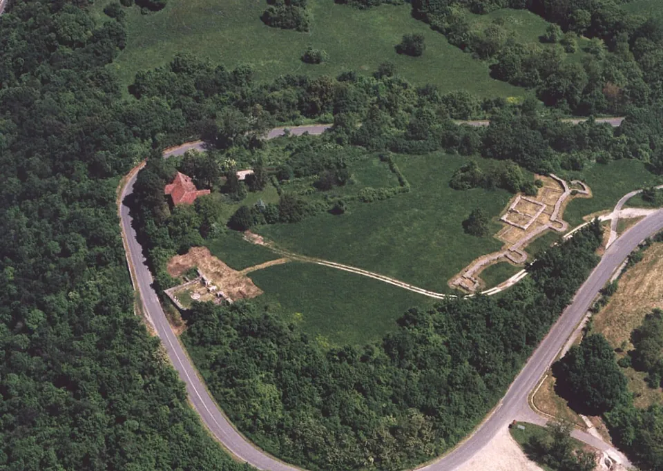 An aerial view of a hilltop meadow ringed by woodland and a looping road, with the pale outlines of excavated walls along its edge, a path across the grass and a red-roofed house among trees.