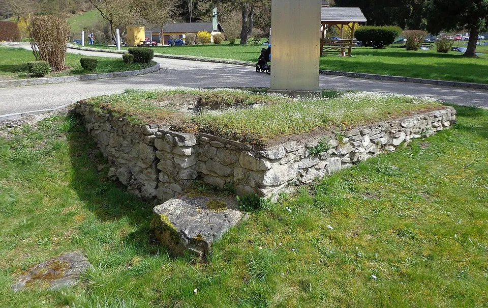 A low square block of rough grey stone masonry, its top grown with grass, beside a lane, with lawns, a parked car and a small timber shelter behind.