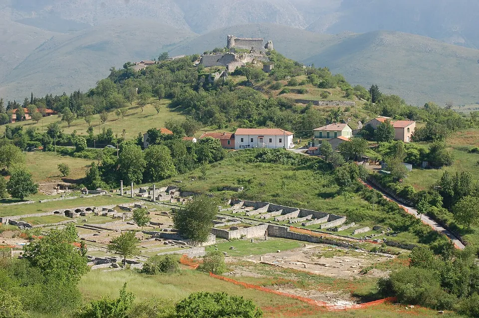 Looking down on a green valley floor where the low grey walls of an excavated town are laid out in a grid, with a few farmhouses beyond and a wooded hill rising behind, a ruined castle tower on its summit and mountains in haze at the back.
