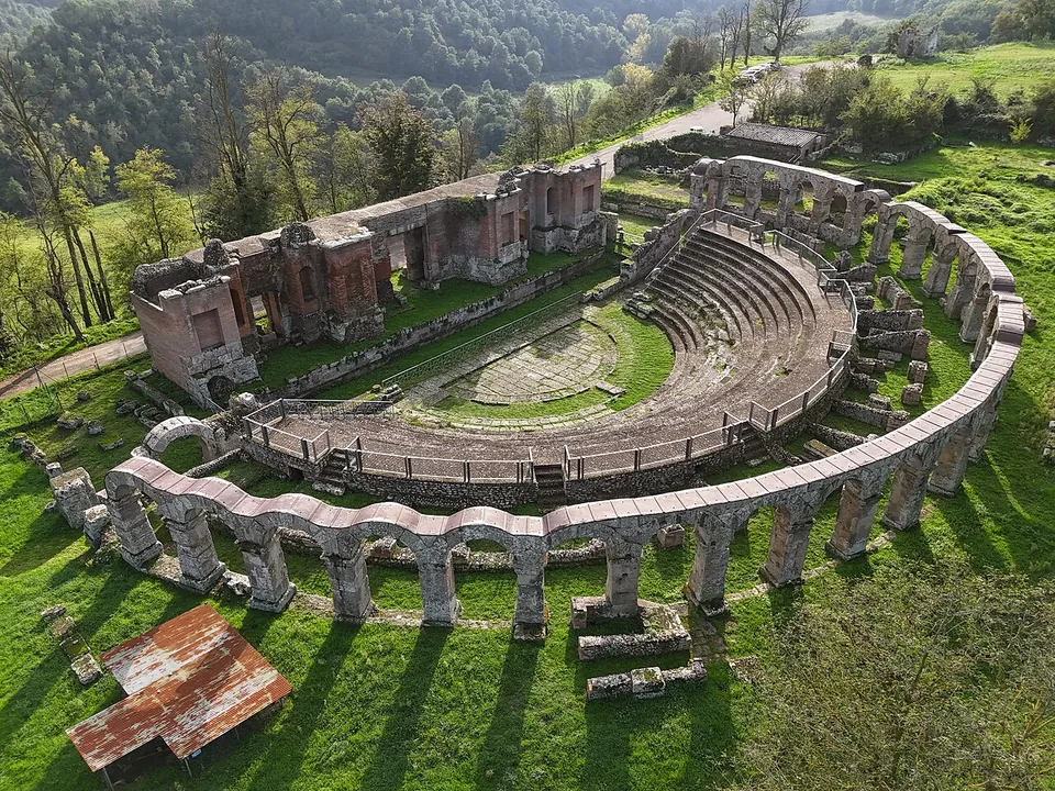 An aerial view of the theatre in a green meadow - a semicircle of stone seating ringed by a curved line of arches, the tall ruined walls of the stage building behind it, a rusty metal roof over one excavated spot in the foreground and wooded slopes beyond.