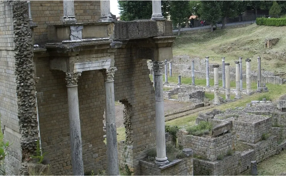 A close view of the standing stage section, two storeys of smooth column shafts with carved capitals set in front of a wall of squared stone, with a row of column stumps on the grass behind.