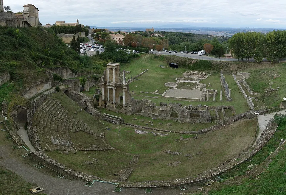 The whole theatre from high above on an overcast day, the half-circle of seating set into a wooded slope, the standing stage section and the excavated ground behind it, with a road, a car park and open countryside stretching to the horizon.