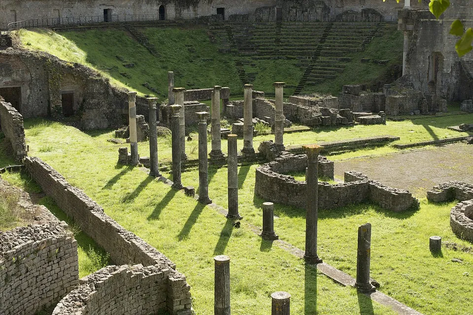 A line of standing column shafts of different heights on a lawn, low stone walls and a curved wall in front of them, and the stone seating rising up the hillside in the background.