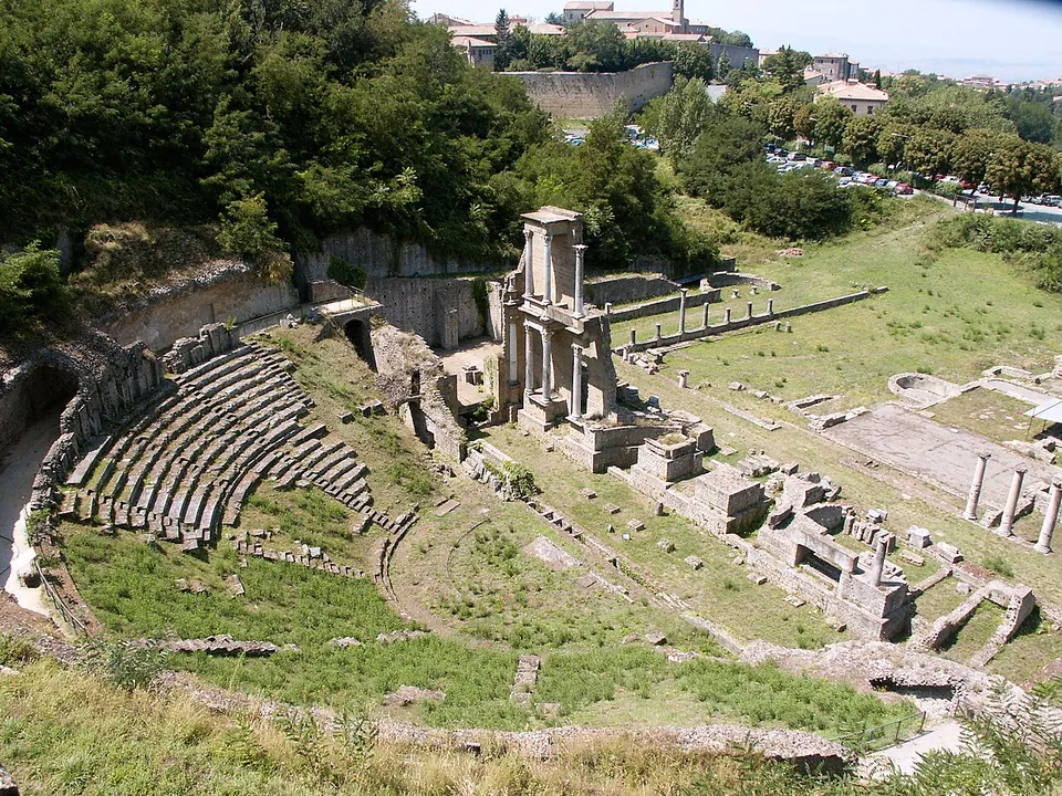 A Roman theatre seen from above, its curved tiers of stone seats set into a grassy hillside, with a tall two-storey fragment of the columned stage front standing beyond them and low walls and column stumps spread over the level ground behind.