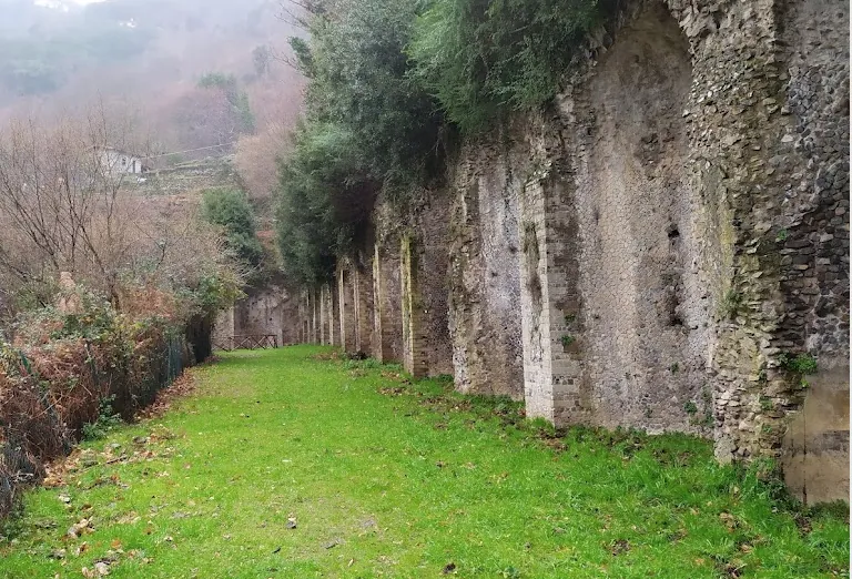A high masonry wall, broken into a long sequence of piers and recesses, runs beside a strip of mown grass, with small trees growing from its top and a misty hillside beyond.