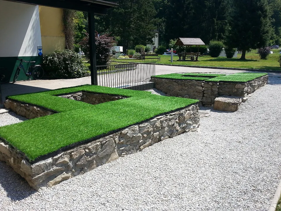 Low stone walls in the outline of a gate tower, topped with artificial turf, set in white gravel beside a building, with lawns and trees beyond.