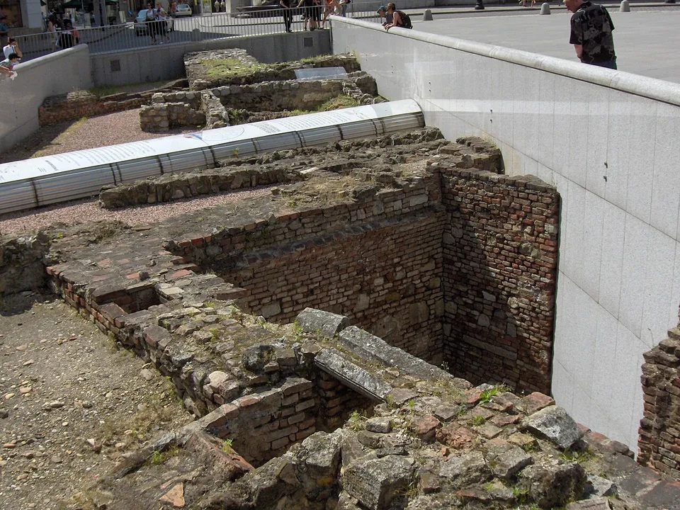 A sunken rectangle in a city square lined with smooth white stone walls, inside it low walls of rubble and brick and a strip of white corrugated roofing, with people at the parapet above.
