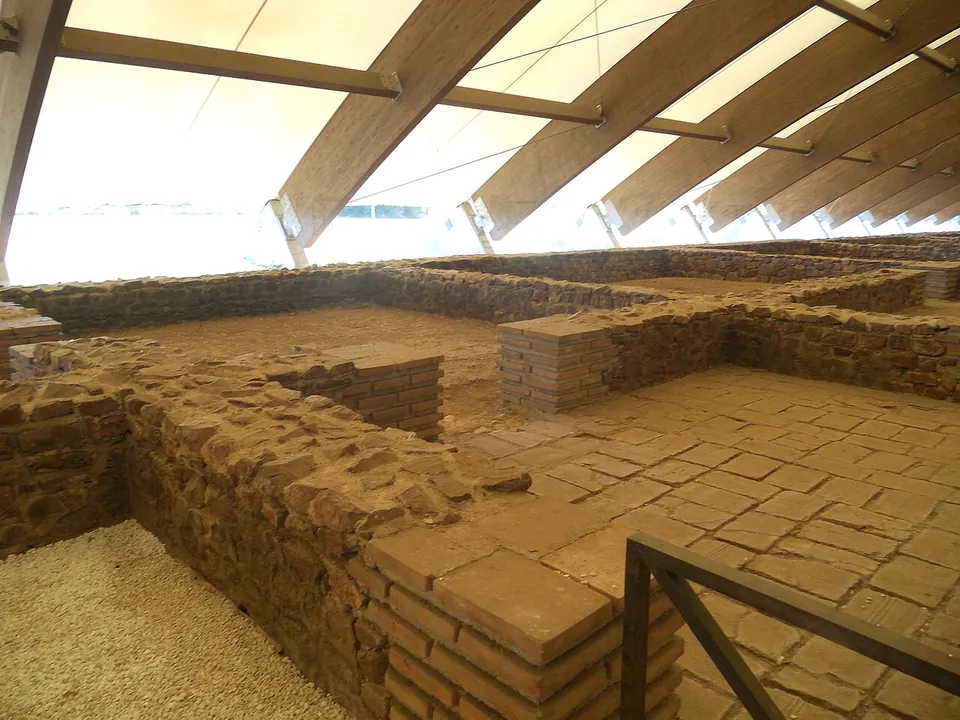 Excavated walls of stone and thin red bricks and a floor of square brick tiles, under a modern canopy of slanting timber ribs and pale fabric, with a metal railing in front.