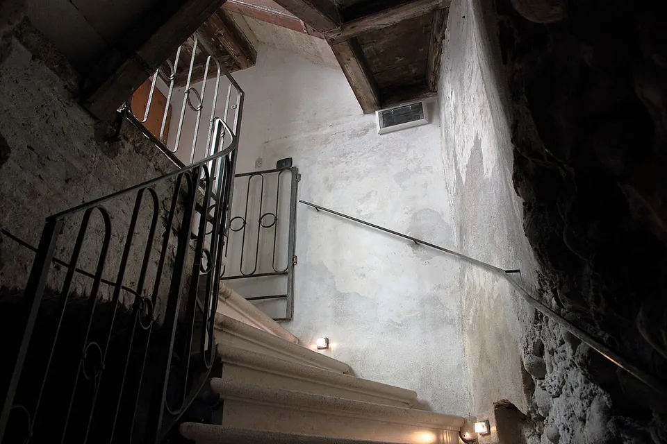 A steep, narrow stone stair turning downwards between whitewashed walls, with an iron railing, a handrail and small lamps at step level.