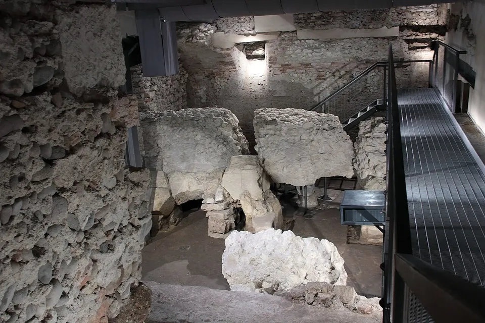 A dim underground excavation seen from above, with walls of rough rubble masonry, large pale blocks of broken masonry lying on an earth floor, and a metal walkway with steps running along the right-hand side.