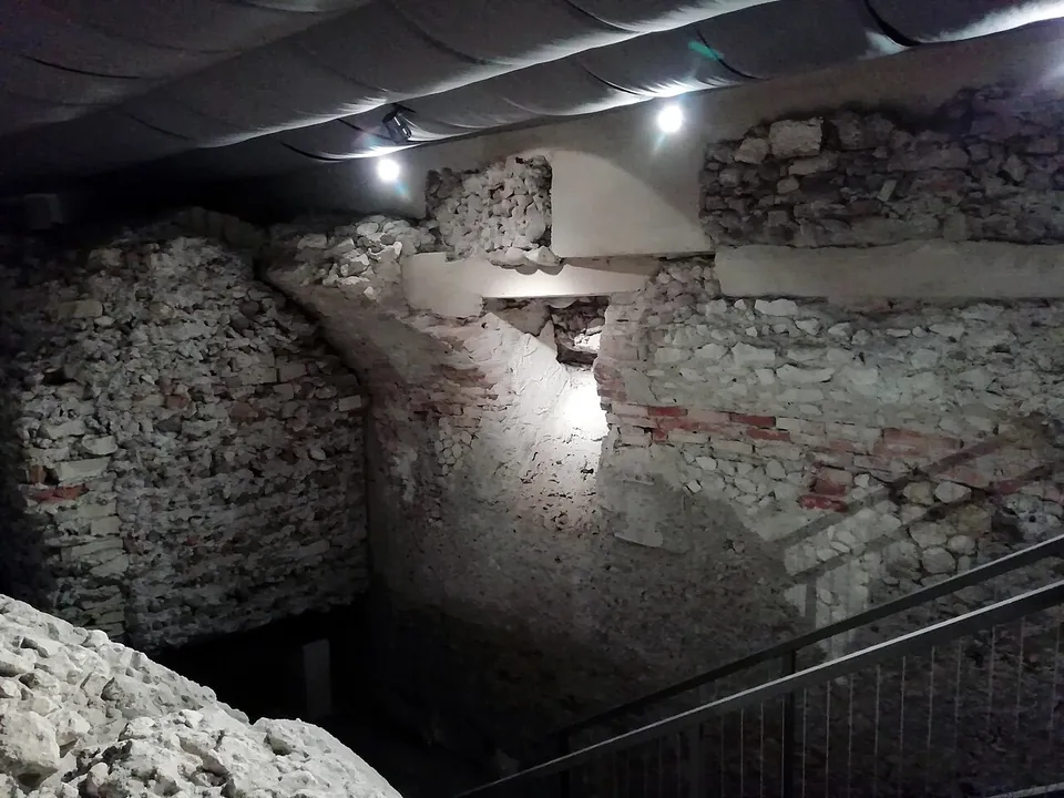 A wall of rough stone with courses of red brick, lit from above, with a sloping, funnel-shaped opening cut through it below a modern fabric ceiling; a stair railing crosses the foreground.