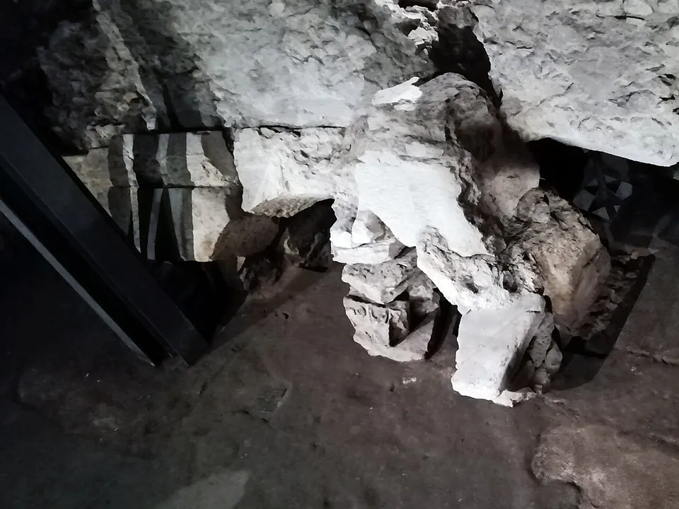 Broken blocks of pale stone and masonry heaped on an earth floor beneath a larger mass of rubble, next to a black steel frame.