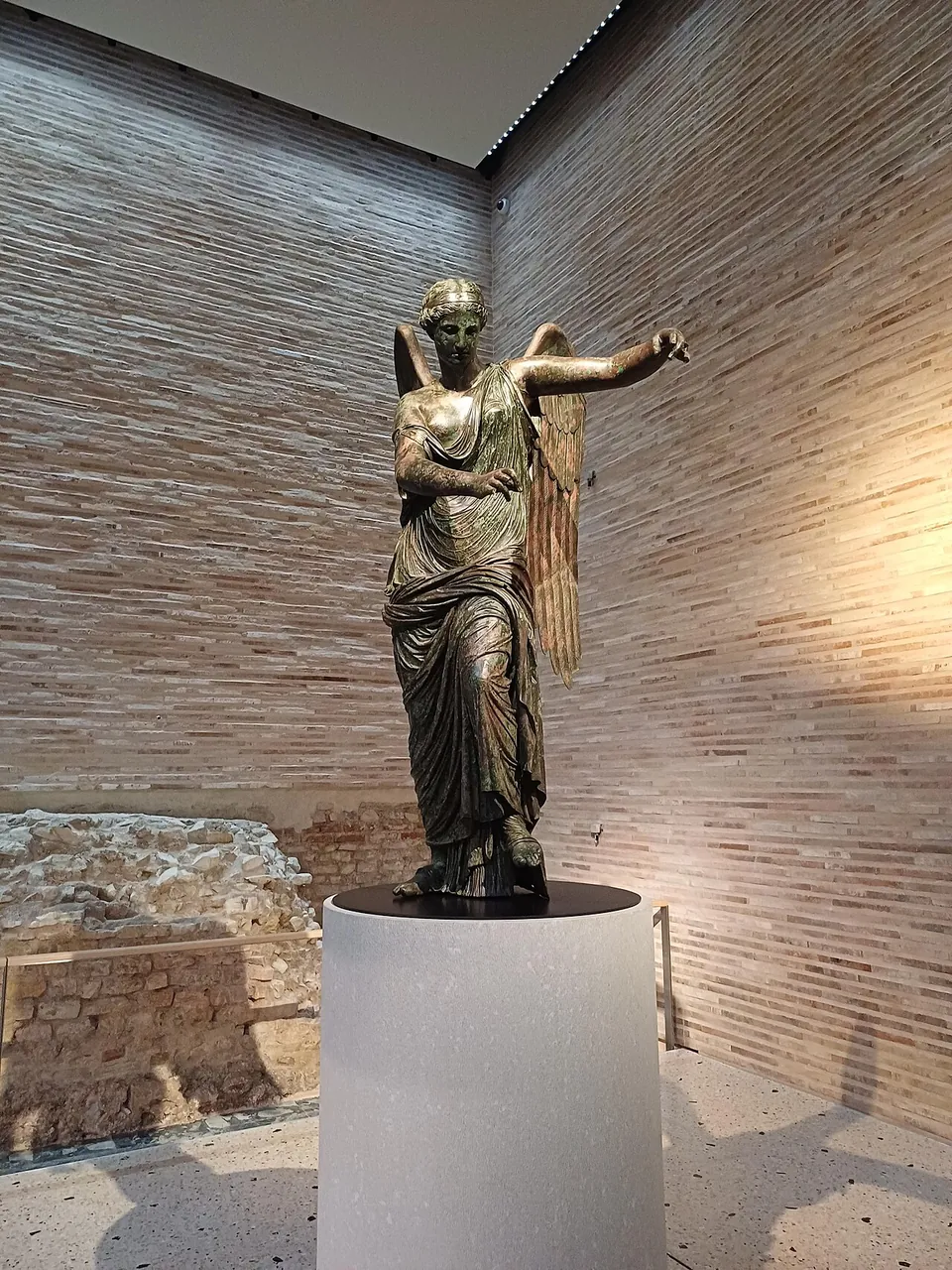 A dark bronze statue of a winged woman in a long draped robe, her arms held out before her as if holding something now missing, standing on a white round pedestal in a tall room with walls of pale brick.