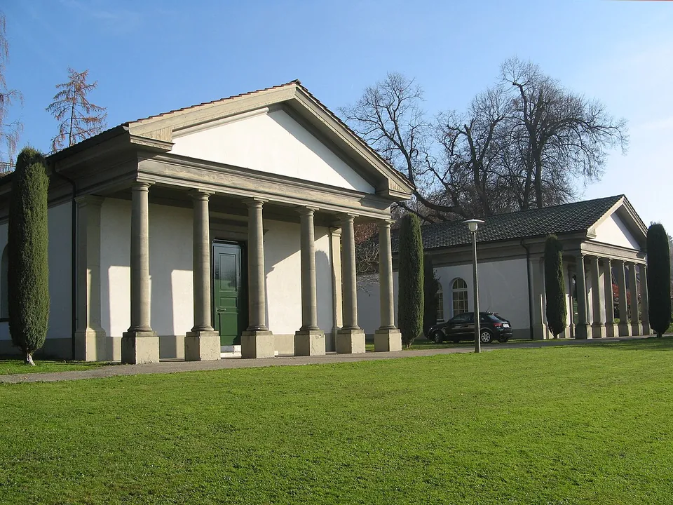 Two small classical buildings with columned porches and pediments, painted white, standing on a lawn among trees and cypresses.