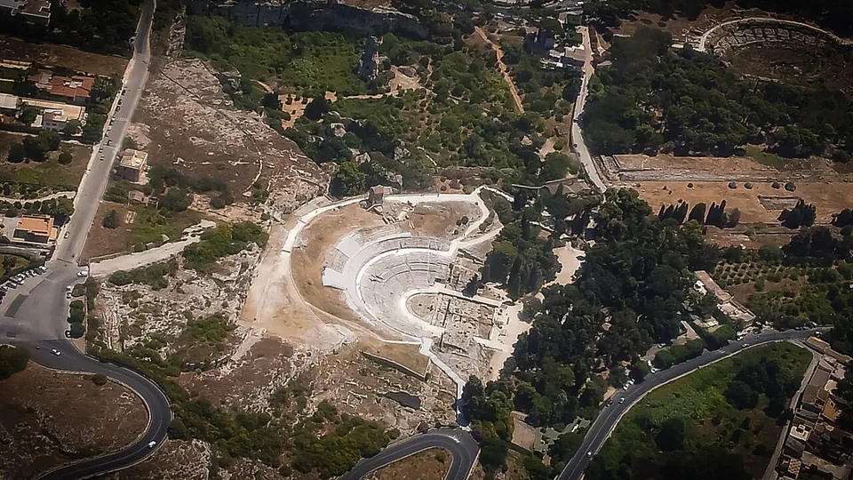 An aerial view of a wooded, rocky hillside cut by roads, with the pale semicircle of an ancient theatre carved into the slope in the centre and a smaller oval arena among trees near the top right.