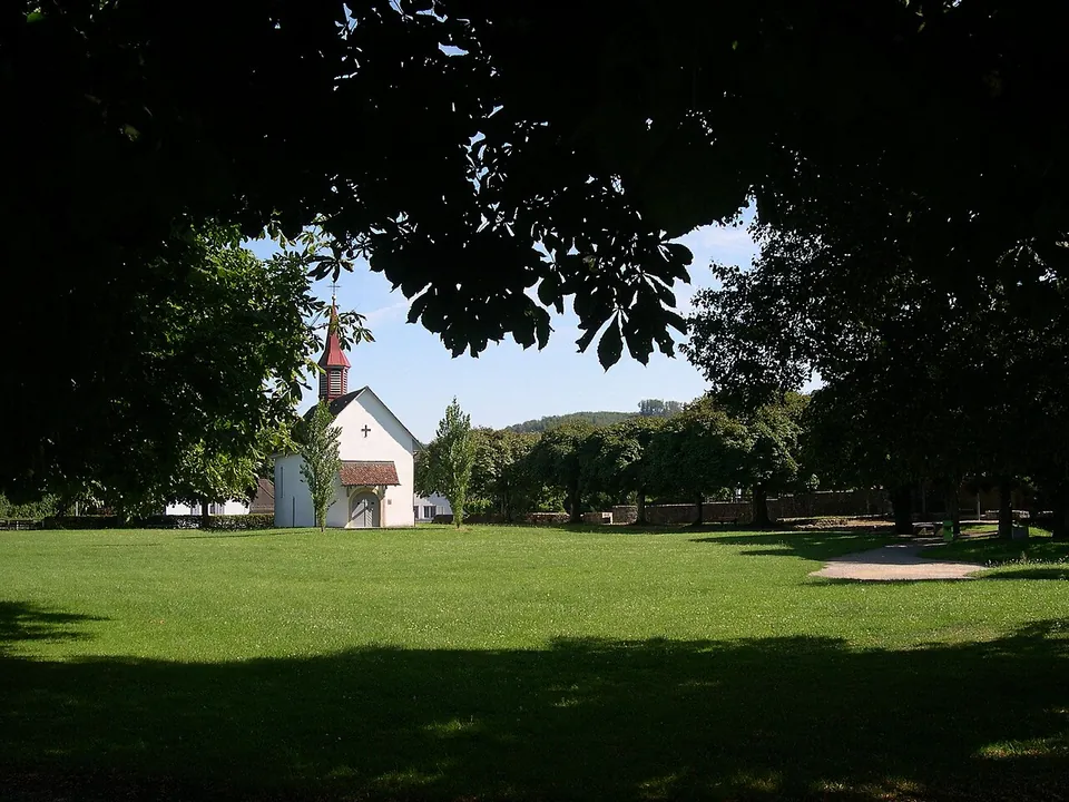 A broad lawn under shady trees with a small white chapel with a red-roofed bell turret on its far side.